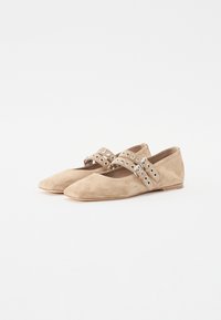 Beige suedesko flate sko med spiss tå, med to brede stropper pyntet med runde metallakenter. Myk tekstur og lav hæl.