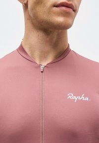 Κοντινό πλάνο ενός ατόμου που φοράει ροζ ποδηλατική φανέλα με φερμουάρ και το λογότυπο «Rapha» στο στήθος και ένα μικρό κρίκο σκουλαρίκι στο αριστερό αυτί.