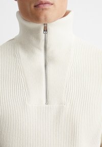 Cremefarbener gerippter Strickpullover mit hohem Kragen, der einen halben Reißverschluss am Hals und ein strukturiertes Steppmuster aufweist. Weicher Stoff.