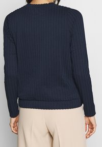 Marinblå ribbad tröja med långa ärmar, texturerat tyg, rundad halsringning och elastisk nederkant, i kombination med ljusbeiga byxor.
