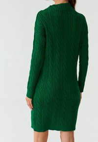 Donna con indosso un vestito in maglia verde a maniche lunghe, vista di spalle, appoggiata a uno sfondo bianco semplice.
