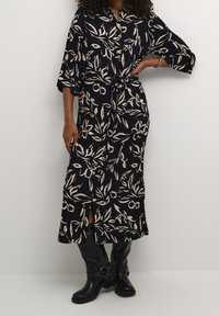 Pitkä musta mekko, jossa vaalea beige kukkakuosi, kauluksellinen pääntie, puolipitkät hihat ja vyötärölle solmittu nauha sekä sivuhalkiot.