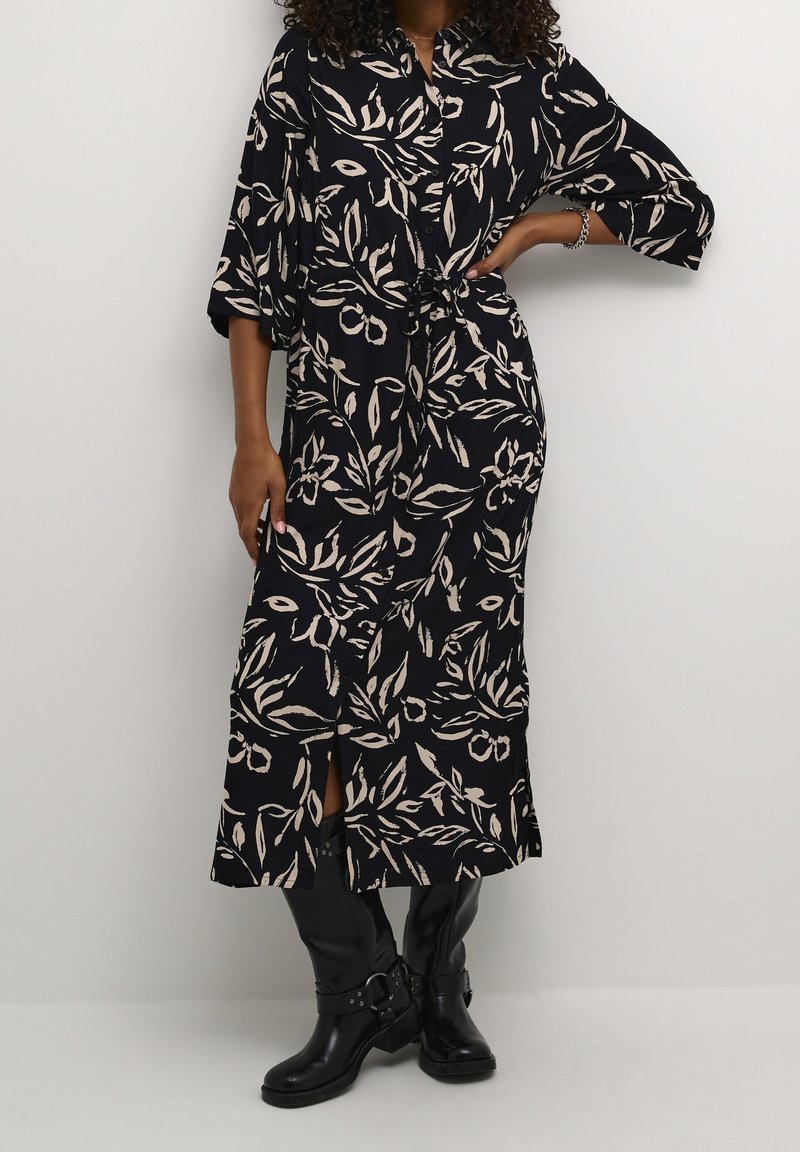 Pitkä musta mekko, jossa vaalea beige kukkakuosi, kauluksellinen pääntie, puolipitkät hihat ja vyötärölle solmittu nauha sekä sivuhalkiot.