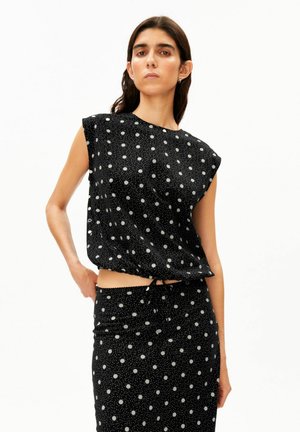 Femme portant un haut noir sans manches et une jupe assortie à pois blancs, debout avec une main sur la hanche sur un fond blanc.