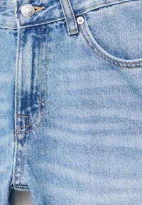 Jean en denim bleu clair en gros plan montrant la poche avant, la boucle de ceinture, le bouton métallique avec gravure de la marque et les détails de couture orange.