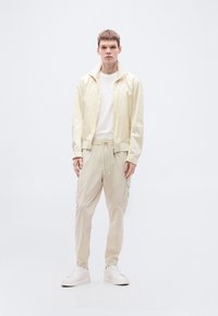 Ανοιχτό μπεζ μπουφάν με φερμουάρ και υφασμάτινο σχέδιο, συνδυασμένο με αντίστοιχο παντελόνι cargo. Άσπρο μπλουζάκι από κάτω, ολοκληρωμένο με άσπρα αθλητικά παπούτσια.