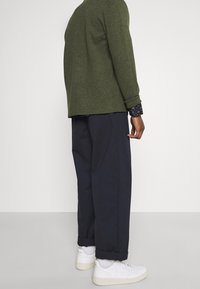 Man i olivgrön jacka, marinblå vida byxor med manschetter, vita strumpor och vita sneakers, stående mot en grönskande ljus vägg.