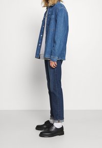 Chaqueta de mezclilla de corte relajado, con cierre de botones y bolsillos en el pecho, superpuesta sobre un sudadera gris. Combinada con jeans ajustados azules y zapatos negros.