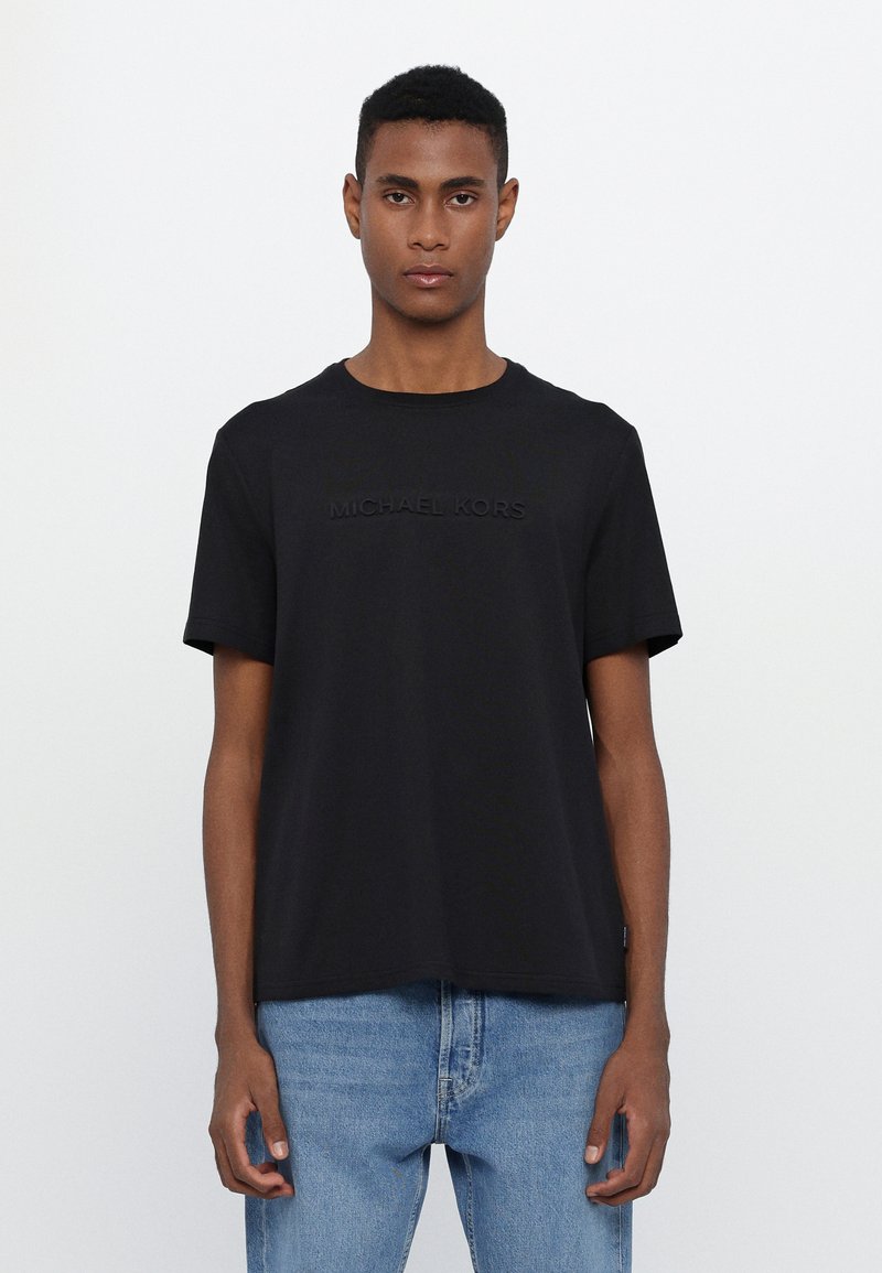 Camiseta de algodón negra con mangas cortas, que presenta el logo en relieve "MICHAEL KORS" en la parte delantera. Forma rectangular con cuello redondo.