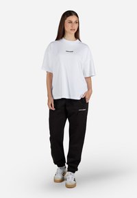 Tricou alb supradimensionat cu mâneci scurte și text negru, asortat cu pantaloni de trening negri cu un mic logo și teniși albi.