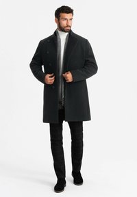 Abrigo largo de lana negra con cuello de muesca y cinco botones, llevado sobre un suéter blanco de cuello alto y una bufanda de cuadros gris, combinado con pantalones de cuadros negros.