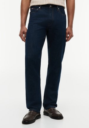 Mann trägt dunkelblaue Jeans mit geradem Bein, braune Lederloafers und einen beigen gestrickten Pullover, der in die Hose gesteckt ist, und steht vor einem weißen Hintergrund.