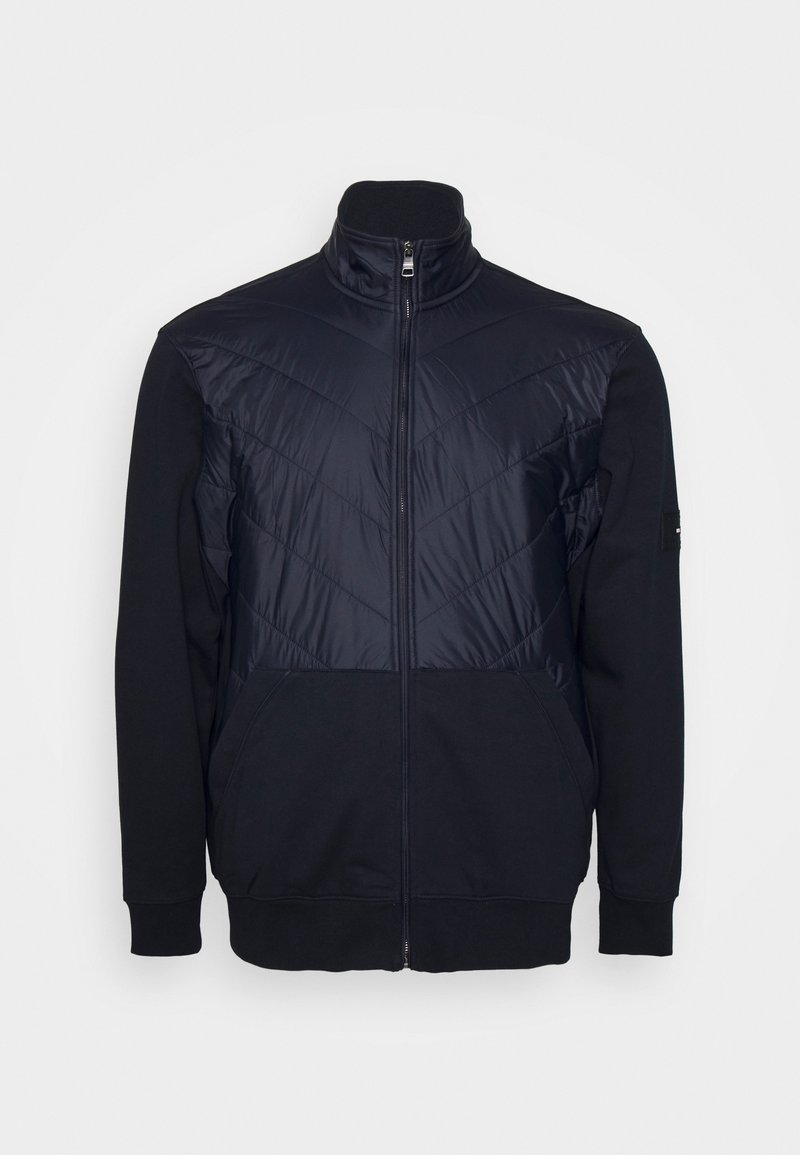 Navy zip-up jacket with a padded front featuring chevron stitching, fleece sleeves, high collar, and side pockets. Textured fabric contrasts design.