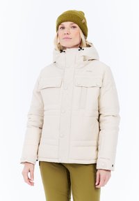 Vrouw in een beige winterjas met capuchon en voorzakken, gecombineerd met olijfgroene broek en een olijfgroene gebreide muts.