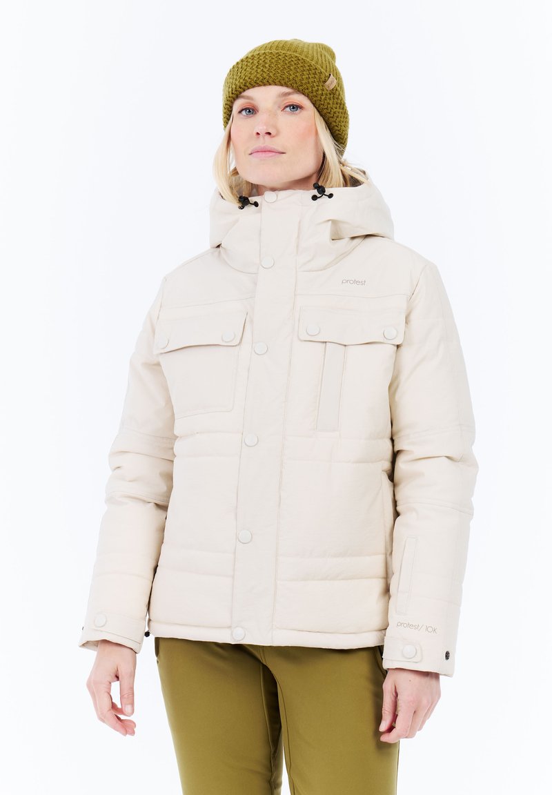 Vrouw in een beige winterjas met capuchon en voorzakken, gecombineerd met olijfgroene broek en een olijfgroene gebreide muts.