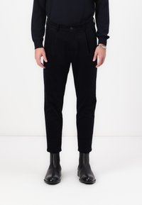 Black corduroy trousers with a tapered leg, pleated front, and elastic waistband, paired with black leather ankle boots.