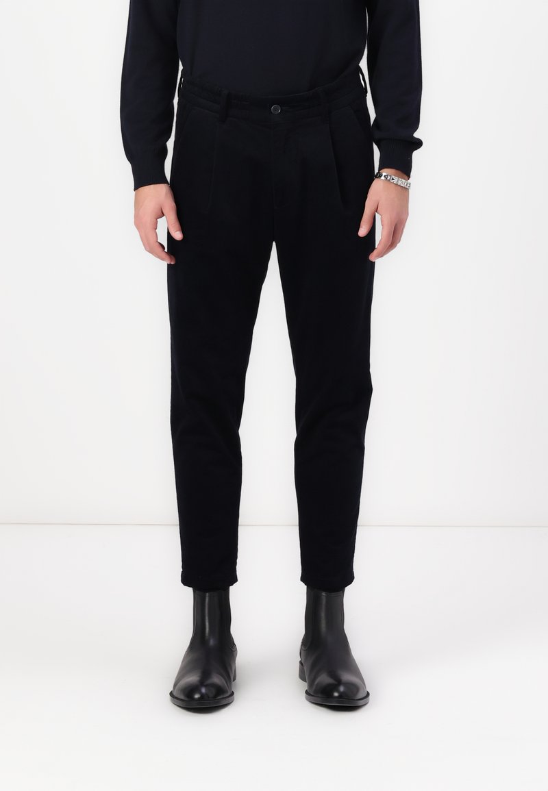 Black corduroy trousers with a tapered leg, pleated front, and elastic waistband, paired with black leather ankle boots.