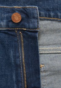 Zbliżenie na materiał dżinsowy z brązowym guzikiem, złotym zamkiem błyskawicznym oraz kontrastującymi teksturami ciemnoniebieskiego i jasnoniebieskiego materiału.