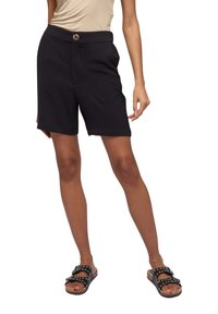 Pantaloni corti neri in tessuto intrecciato con chiusura a bottone, tasche laterali e vestibilità rilassata. Abbinati a sandali con borchie neri.