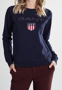 Granatowa bluza z nadrukiem logo "GANT" i tarczą z czerwono-białymi paskami; ściągacz na dekolcie i mankietach; miękki materiał.