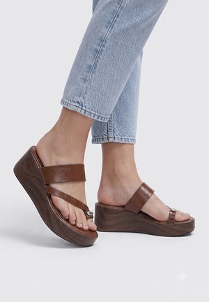 Pies con sandalias marrones de cuña con plataforma y dos tiras de cuero, combinados con vaqueros cortos azul claro sobre un fondo liso.