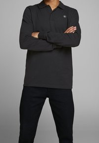 Svart långärmad polo med krage, prydd med en liten rund logotyp på vänster bröst, i kombination med mörka jeans.