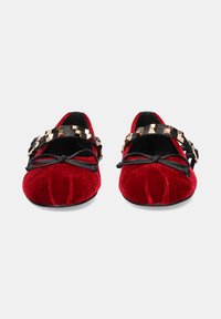 Las bailarinas de terciopelo rojo presentan una punta redondeada, acentos de lazo negro y una correa decorativa con adornos cuadrados en dorado y negro.