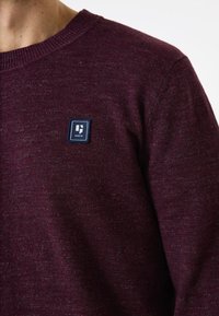 Bordeaux ronde hals trui, textuurstof met een blauwe vierkante patch met het label "Garcia" op de linkerborst, gebreide halslijn en manchetten.