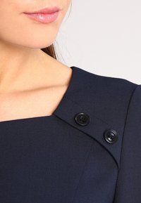 Robe bleu marine avec un décolleté carré, ornée de deux boutons noirs sur les épaules et en tissu texturé.