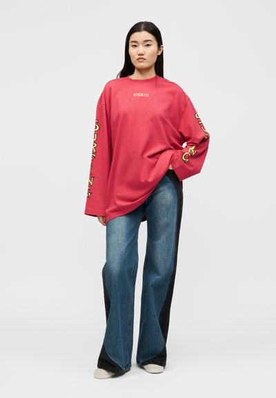 Γυναίκα στέκεται με το χέρι στην τσέπη, φορώντας υπερμεγέθη κόκκινο μακρυμάνικο πουκάμισο με κίτρινο κείμενο και φαρδιά μπλε τζιν με μαύρες πλευρικές ραφές.