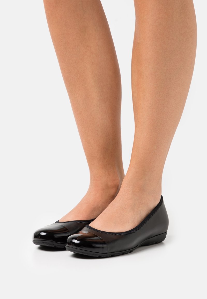 Jambes portant des ballerines noires à petit talon compensé avec des bouts brillants, debout sur un fond clair uni.