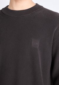 Dark gray ribbed sweater with a round neckline, featuring a subtle embossed brand logo on the chest and textured fabric design.