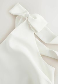 White garment with a large fabric bow tied at the shoulder on a plain light background.