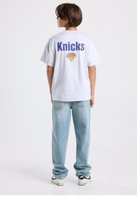 Gray cotton T-shirt featuring "Knicks" in bold blue lettering on the back, paired with light blue baggy jeans and black sneakers.