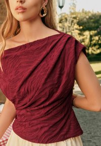 Burgundyröd topp med texturerade, vågiga mönster; off-shoulder design med korta ärmar; matchad med guldhänge.