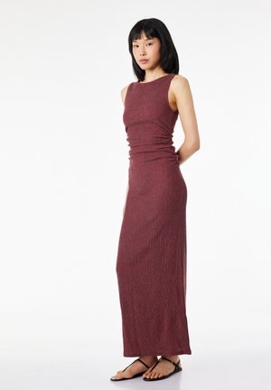 Femme aux cheveux noirs et lisses portant une robe maxi bordeaux sans manches côtelée et des sandales plates noires, debout devant un fond blanc.