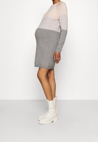 Gravid person iklädd en långärmad, färgblockerad stickad klänning i nyanser av beige och grått, med vita kraftiga stövlar, stående på sidan.