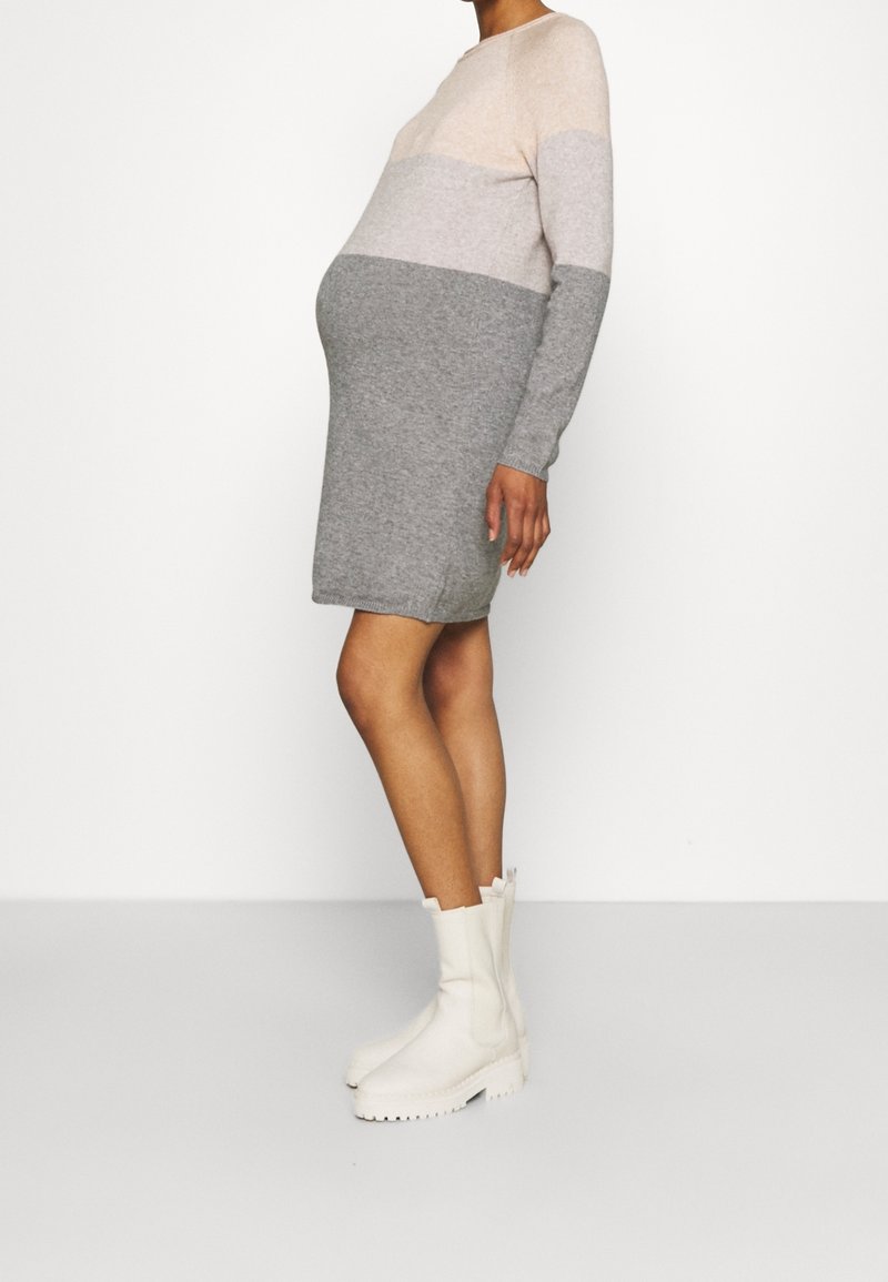 Gravid person iklädd en långärmad, färgblockerad stickad klänning i nyanser av beige och grått, med vita kraftiga stövlar, stående på sidan.