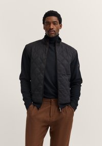 Gilet trapuntato nero con colletto alto, abbinato a un dolcevita nero. Il modello indossa pantaloni marroni. Presenta chiusura con zip e tasche laterali.