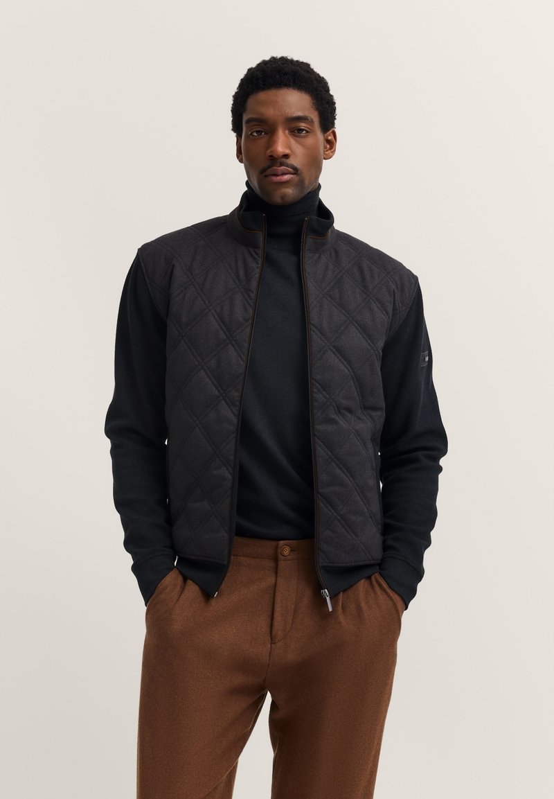 Gilet trapuntato nero con colletto alto, abbinato a un dolcevita nero. Il modello indossa pantaloni marroni. Presenta chiusura con zip e tasche laterali.