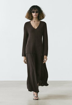 Frau mit lockigem Haar, die eine Sonnenbrille, einen dunkelbraunen V-Ausschnitt-Pullover, einen fließenden dunkelbraunen Rock und schwarze Sandalen trägt und vorwärts geht.