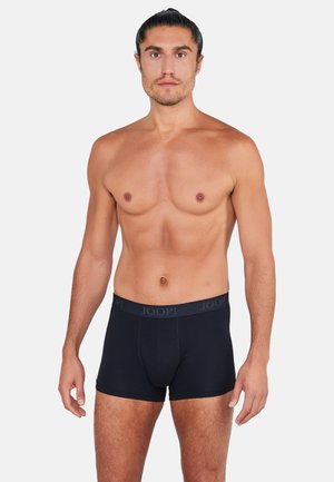 Oberkörperfreier Mann mit zurückgebundenen Haaren trägt schwarze JOOP! Boxershorts und steht vor einem schlichten hellen Hintergrund.