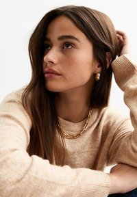 Maglione beige a maglia con polsini a coste, abbinato a una collana e orecchini in oro. Capelli lunghi e lisci, tono di pelle morbido e sfondo neutro.
