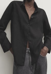 Chemise noire boutonnée en lin, avec une coupe décontractée, des manches longues et un col, présentée avec un pantalon en denim gris.