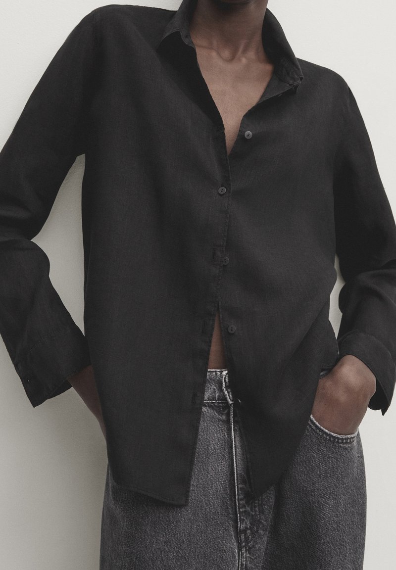 Chemise noire boutonnée en lin, avec une coupe décontractée, des manches longues et un col, présentée avec un pantalon en denim gris.