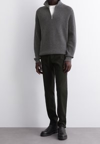 Pull à col zippé côtelé gris, T-shirt blanc en dessous, pantalons vert foncé, bottes noires. Tissu texturé et coupe décontractée.