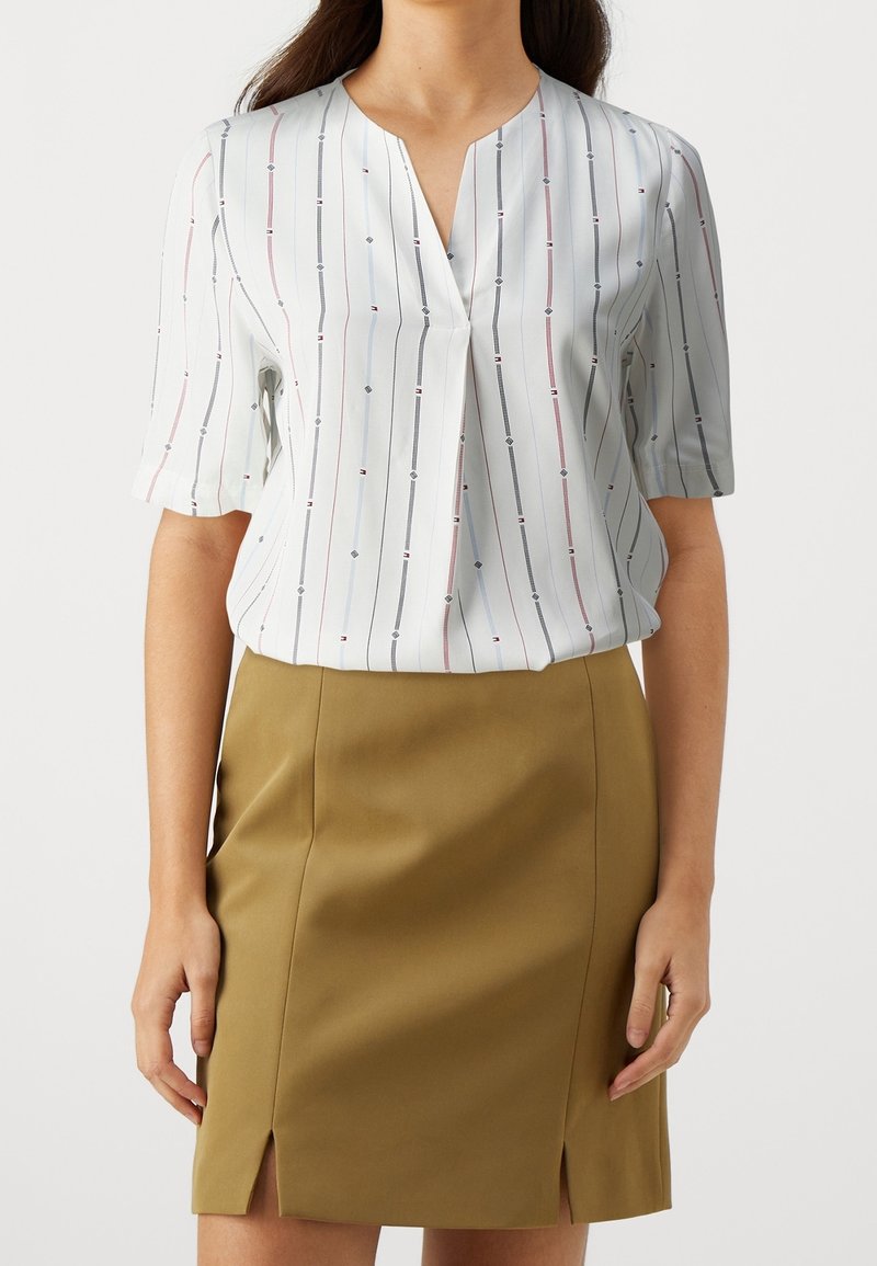 Femme portant un chemisier blanc à manches courtes avec des rayures verticales et une jupe beige longueur genoux avec des fentes à l'avant.