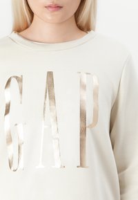 Lähikuva henkilöstä, joka käyttää vaaleanbeigeä hupparia, jossa on suuri metallinhohtoinen kultainen "GAP" -teksti edessä.