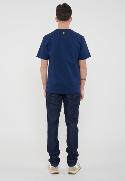 T-shirt di cotone blu navy con maniche corte, scollatura rotonda e un piccolo logo giallo sul retro. Abbinata a jeans in denim scuro e sneakers chiare.