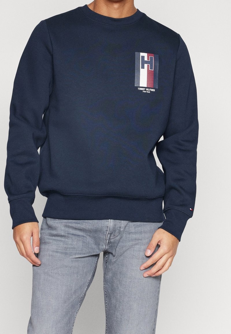 Homme portant un sweat-shirt bleu marine Tommy Hilfiger avec écusson logo sur la poitrine, assorti à un jean gris clair, debout devant un fond uni.